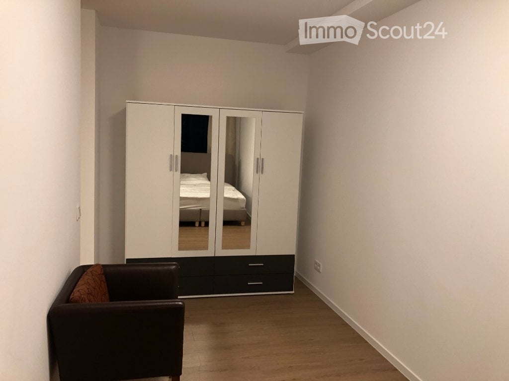 A modern, minimalist bedroom with a large white wardrobe, a black leather chair with a patterned pillow, and a glimpse of a bed in the background.