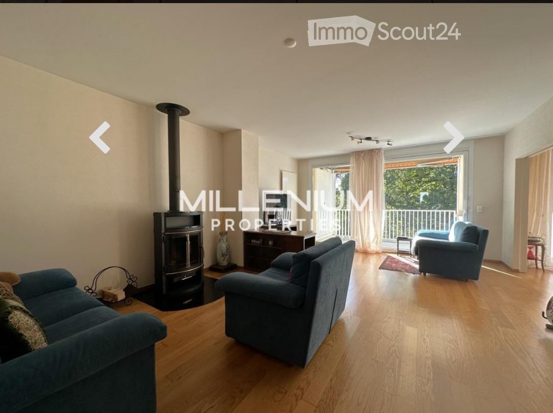 Spacious living room with wooden flooring, fireplace, large windows, and balcony access. Features a blue sofa, armchair, and small table.