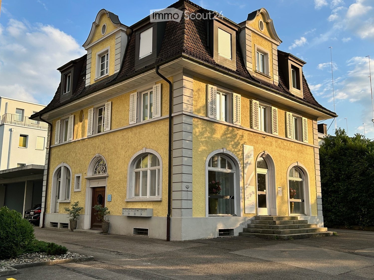 3-story yellow building with brown roof, arched windows, yellow exterior walls, white shutters, steps leading to the entrance, letter boxes, potted plants in front, garage on the left, parking lot in front, trees, bushes, cars parked on the street.