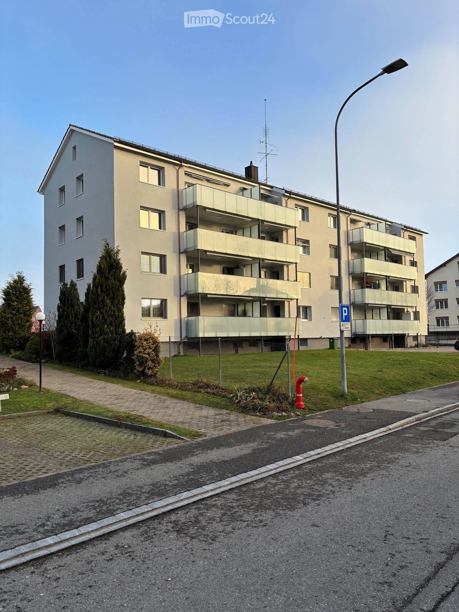 Four-story residential building with multiple balconies, surrounded by greenery and a paved walkway.