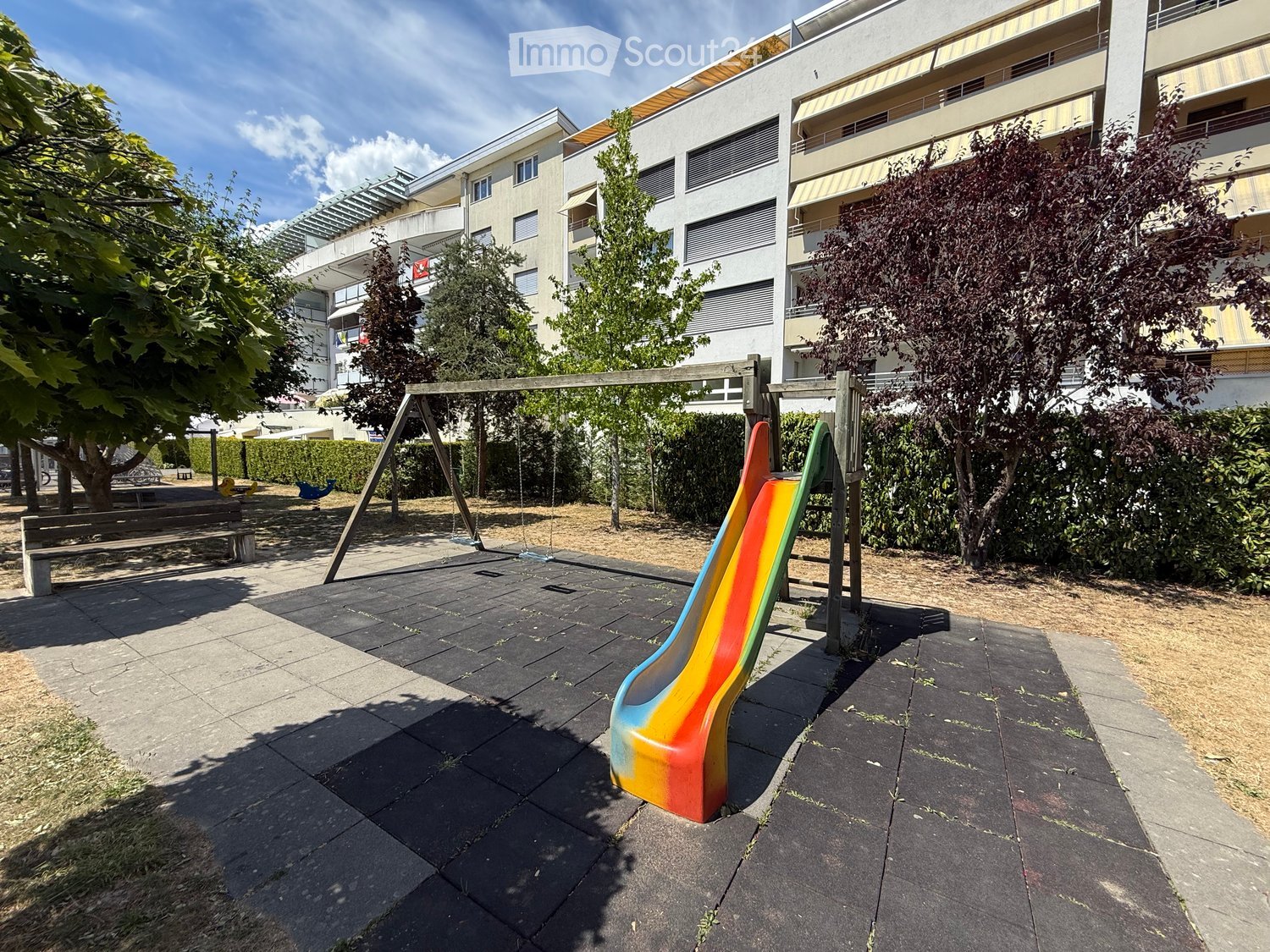 Playground area with a colorful slide, swings, and benches, surrounded by trees and modern apartment buildings.