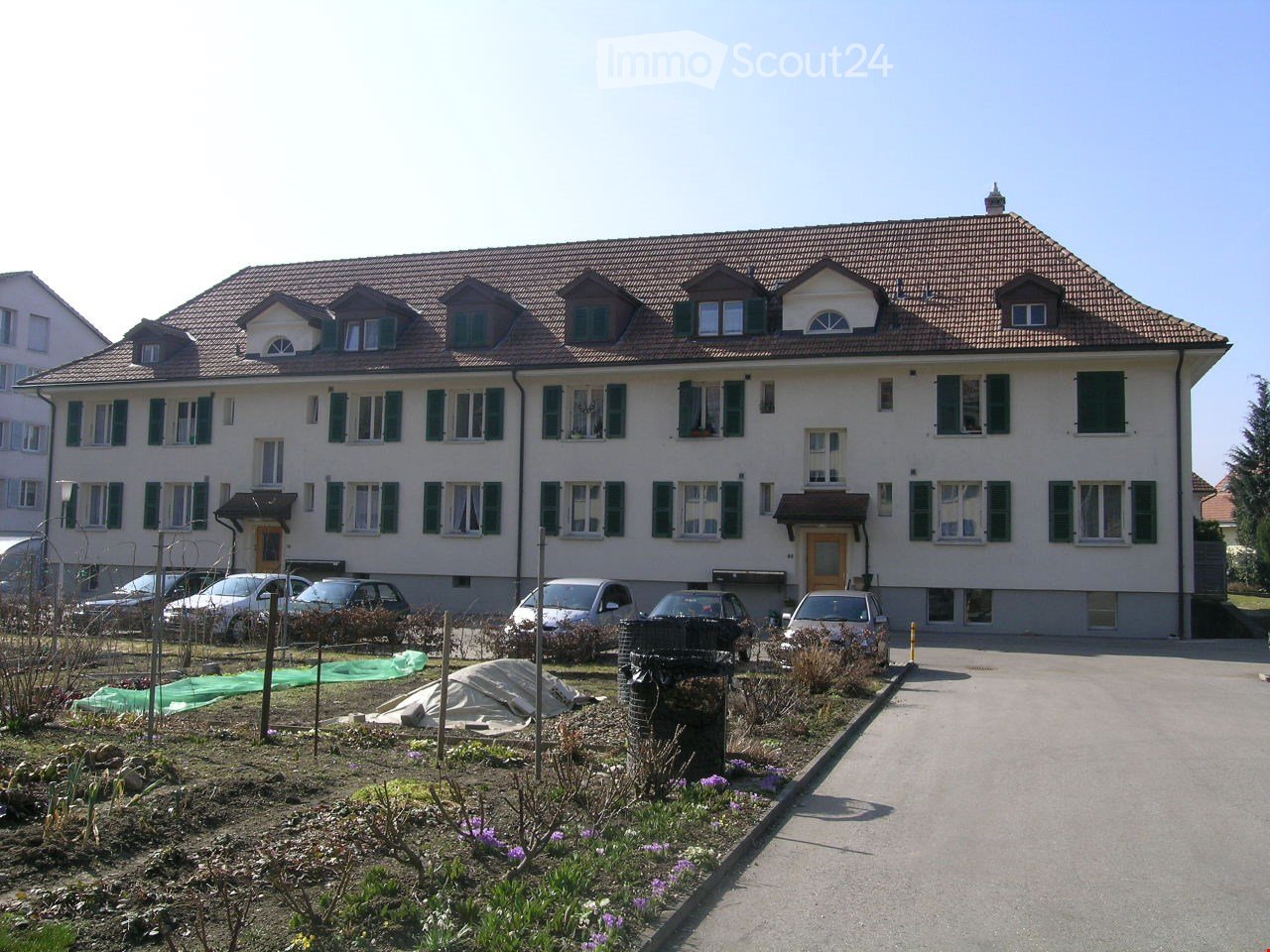 A two-story house with a tiled roof, multiple windows, and a driveway with several parked cars. The front yard has some plants and a small garden area.
