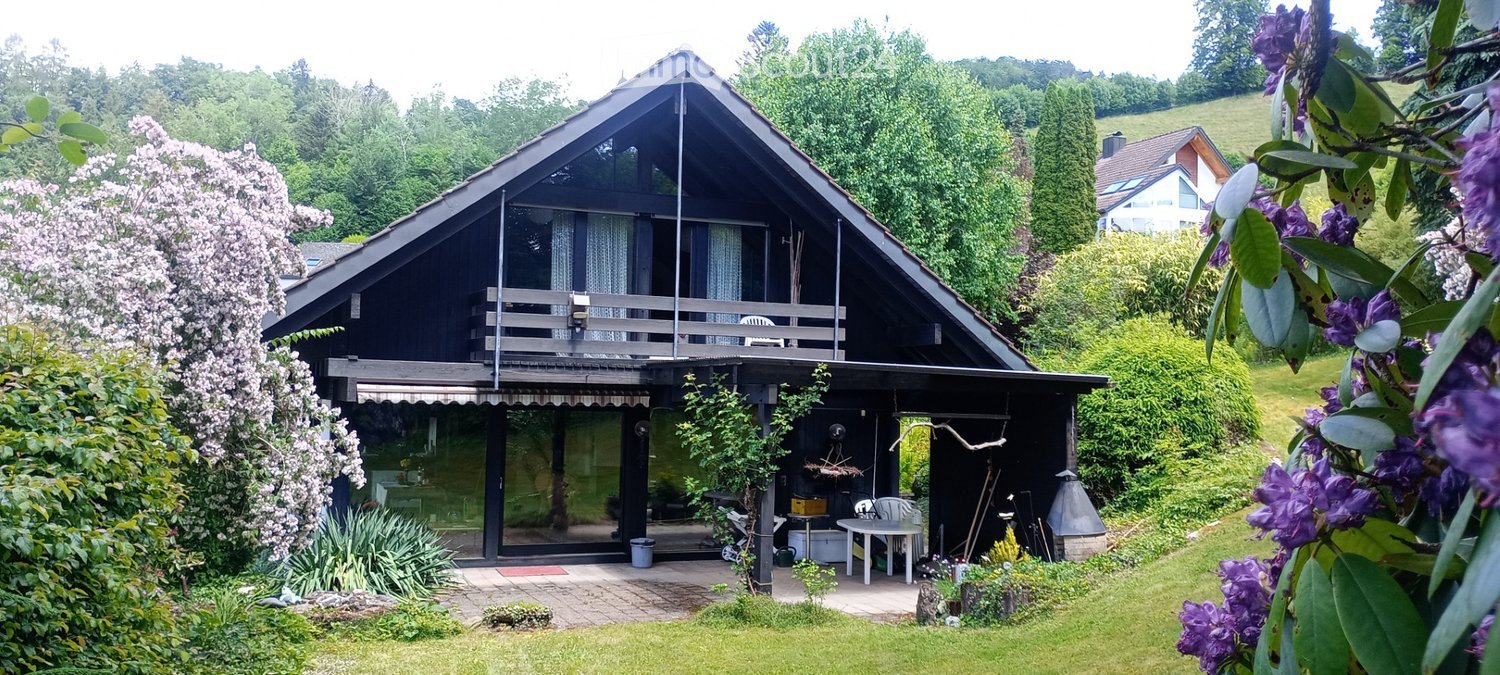 Two-story house with a dark wooden exterior, large windows, and a balcony on the upper level. The surrounding area includes a well-maintained garden with flowering trees and shrubs.