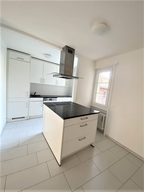 Modern kitchen with white cabinets, a central island with a black countertop, and stainless steel appliances including a stove and range hood. The space features tiled flooring and a window with a radiator below.