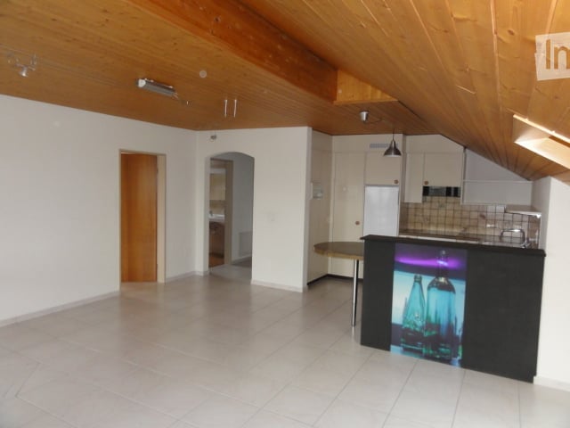 Open-plan kitchen and living area with white tiled flooring, wooden ceiling beams, and white cabinetry. The space is unfurnished, showing only structural elements like walls, doors, and built-in cabinets.