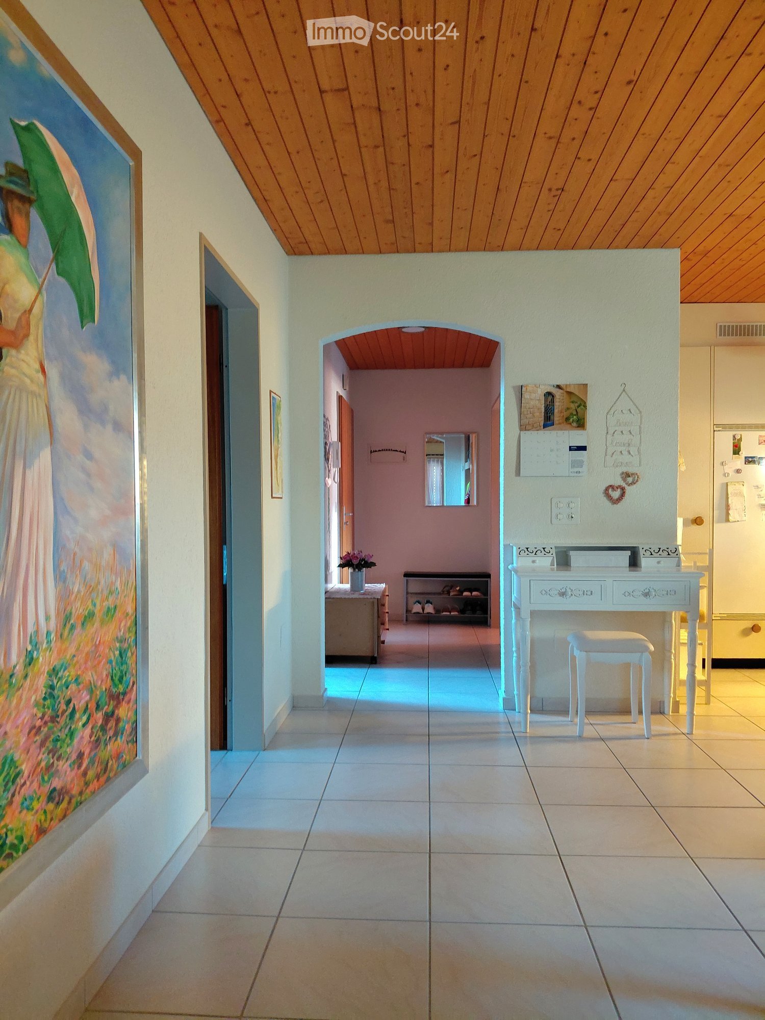 Bright hallway with tiled flooring, wooden ceiling, and white walls. Features a small white desk with a stool, a shoe rack, and a painting on the wall.
