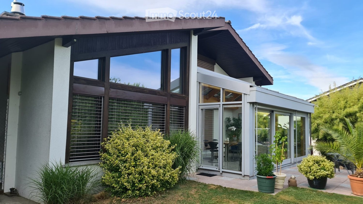 Modern two-story house with large windows, sliding glass doors, and a well-maintained garden with potted plants.