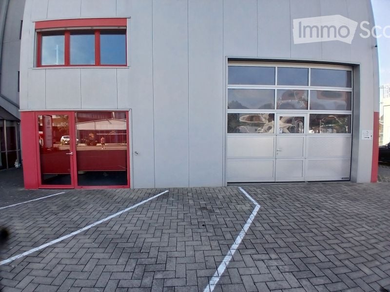 Modern building with a large garage door and a glass entrance. The facade is grey with red trim around the windows and doors. The ground is paved with bricks and marked parking spaces.