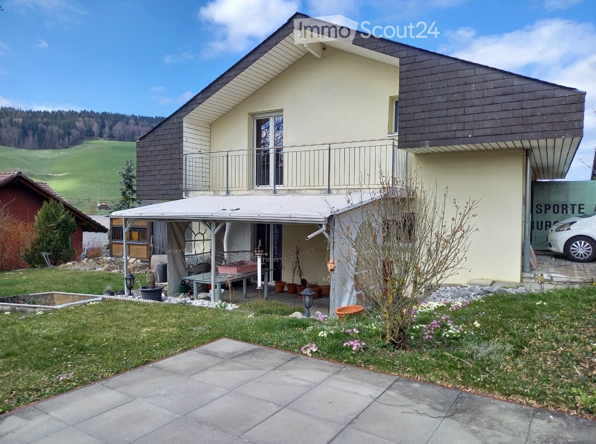 Two-story house with a modern design, featuring a balcony, a covered patio area, and a well-maintained garden with a view of green hills.