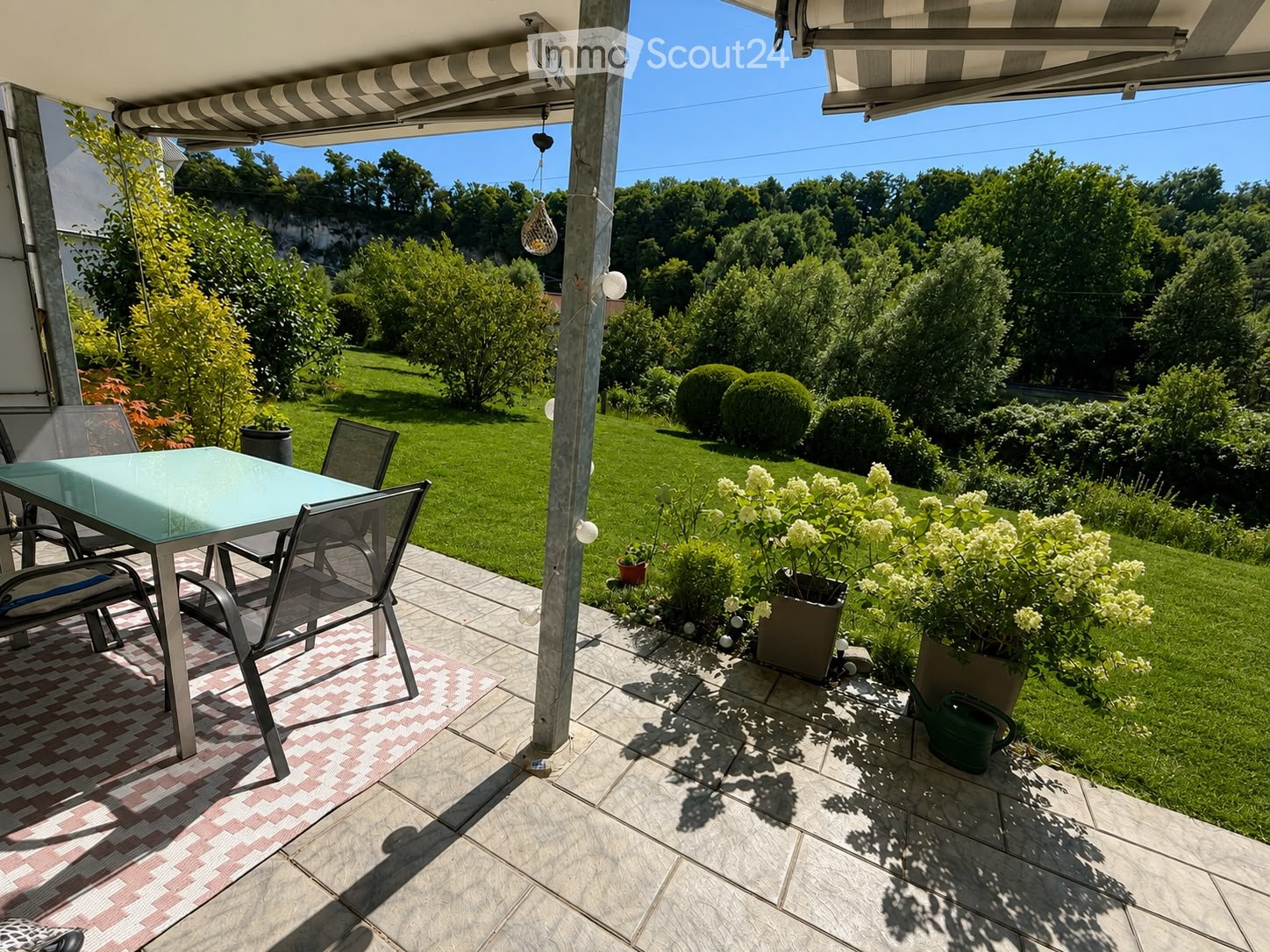 Outdoor patio with a glass table, metal chairs, potted plants, and a well-maintained lawn.