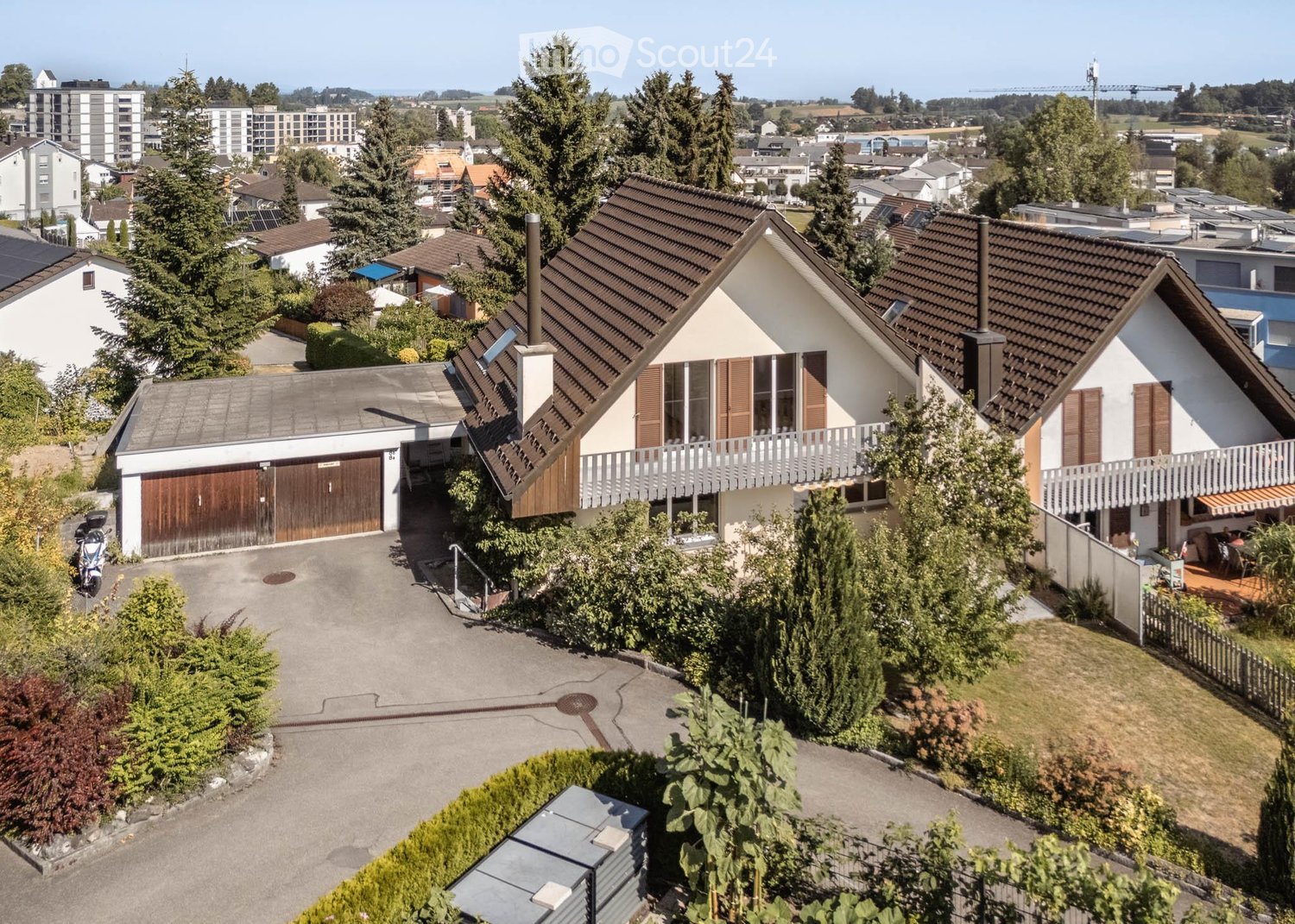 Aerial view of a two-story house with a garage, surrounded by greenery and neighboring houses. The house features a balcony and a well-maintained garden.