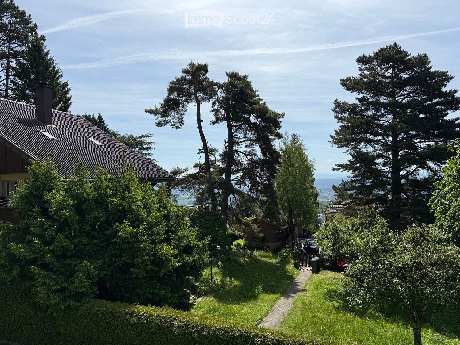 A well-maintained garden with a gravel pathway leading to a house, surrounded by tall trees and greenery, with a view of distant hills.
