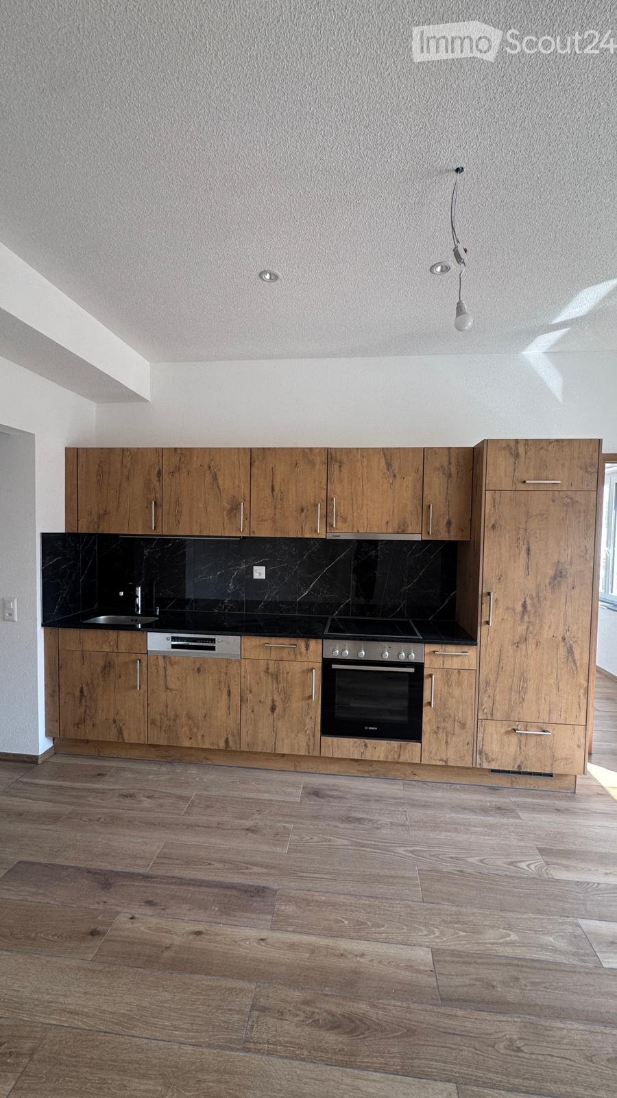 Modern kitchen with wooden cabinets, black countertops, and stainless steel appliances including an oven and dishwasher. The floor is made of light wood.