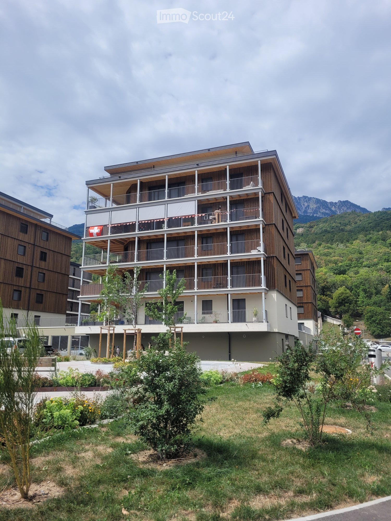 Modern multi-story building with wooden balconies and a Swiss flag, surrounded by greenery and mountains.