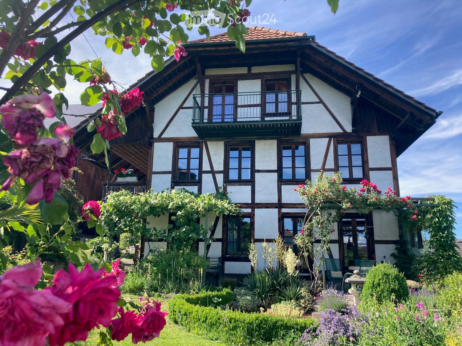 Traditional half-timbered house with white plaster walls, multiple windows, and a balcony on the upper floor, surrounded by a well-maintained garden with various plants and flowers.