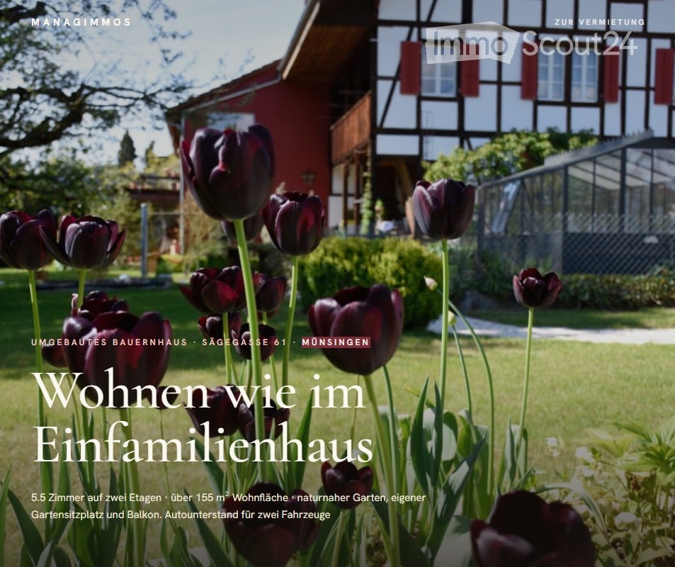 A well-maintained garden with dark red tulips in the foreground, featuring a traditional half-timbered house with red shutters and a greenhouse in the background.
