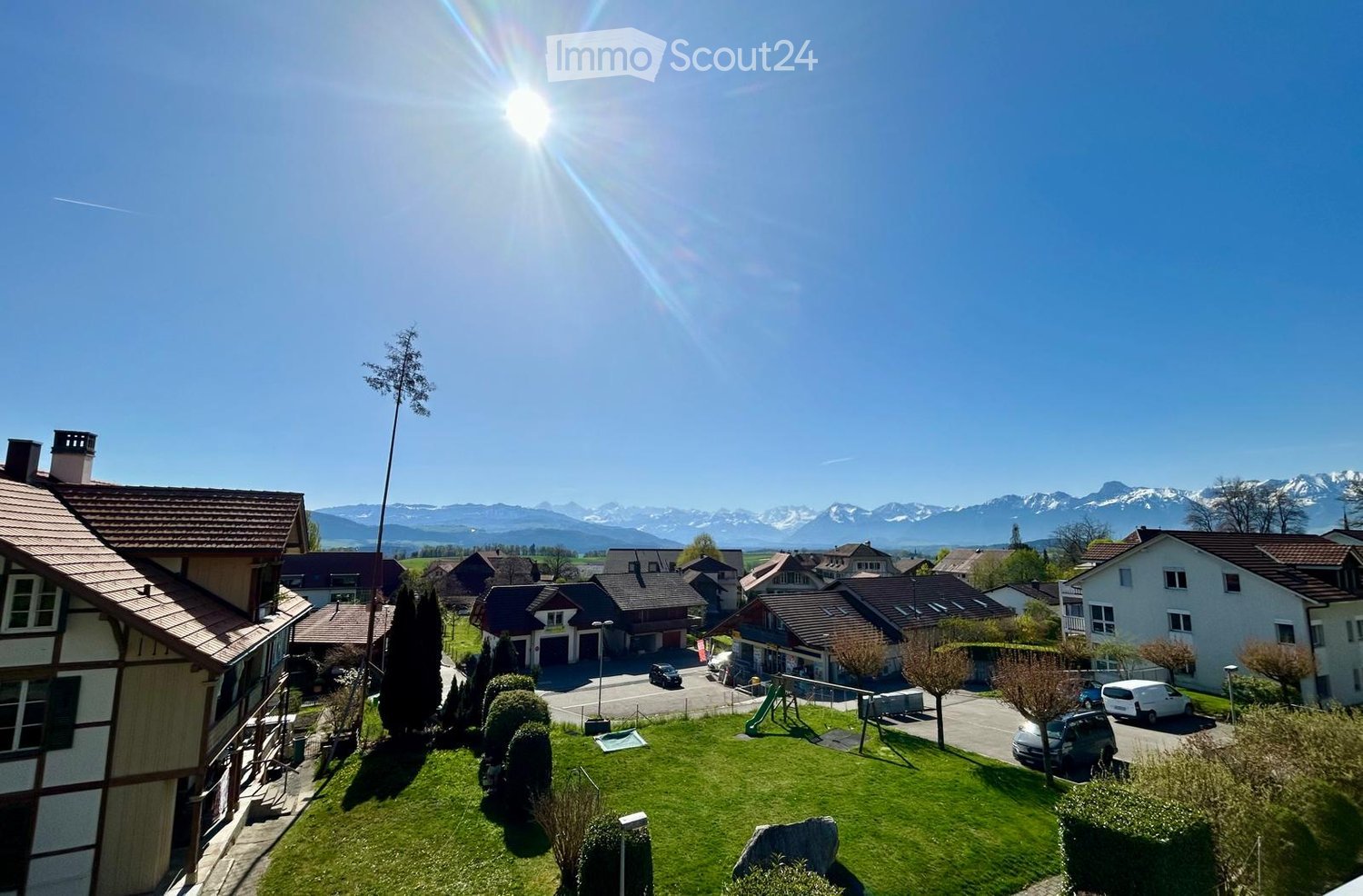 A sunny garden area with green grass, trees, and a playground, overlooking a residential neighborhood with houses and a mountain range in the background.