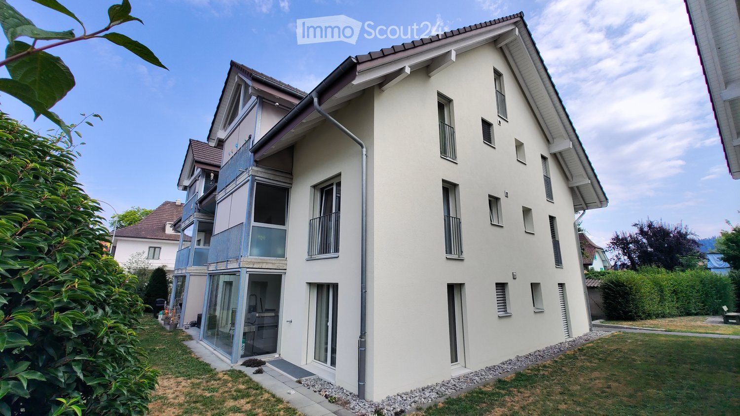 Modern two-story house with white exterior, multiple windows, and balconies. Features a small garden area with a patio and outdoor furniture.