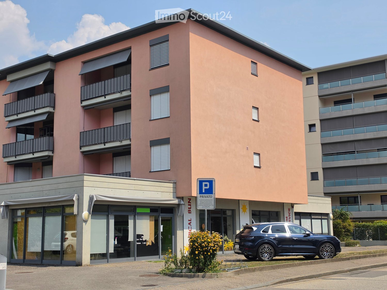Modern multi-story residential building with pink facade, balconies, and private parking area.