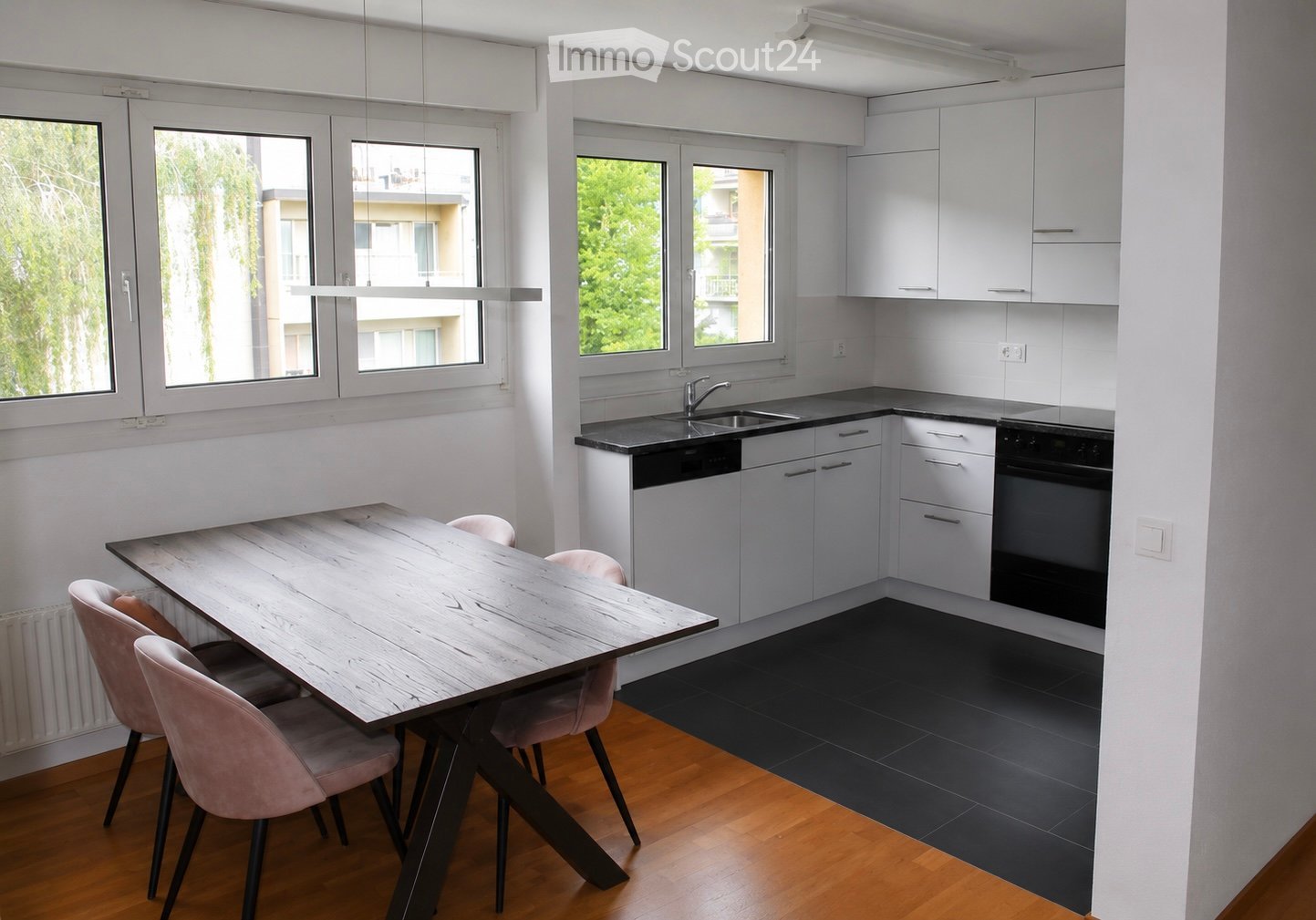 Modern kitchen with white cabinets, black countertops, a wooden dining table with four pink chairs, and large windows offering a view of greenery.