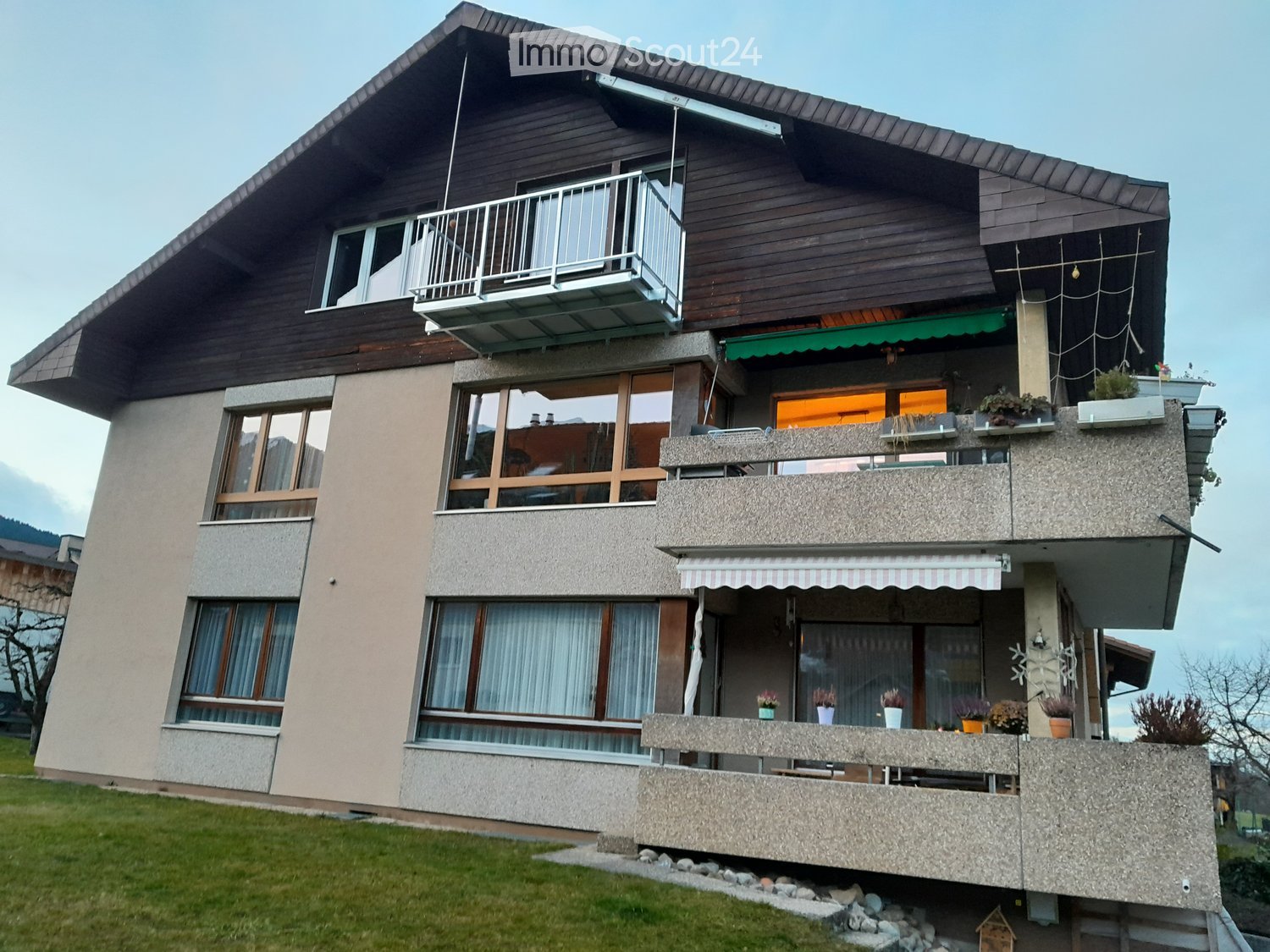 Two-story house with wooden facade, multiple balconies, large windows, and a well-maintained lawn.