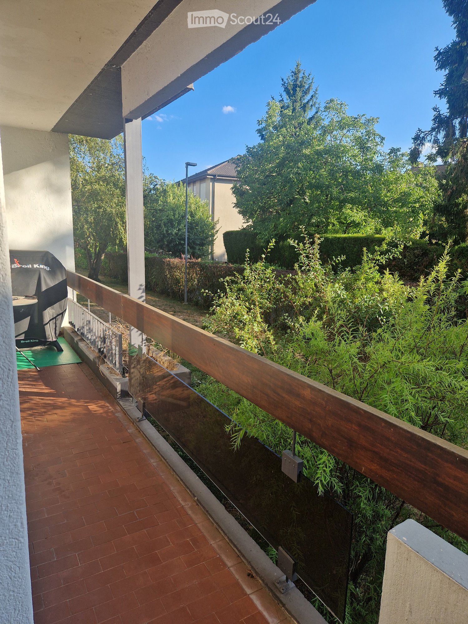 Balcony with wooden railing, tiled floor, and outdoor furniture including a table and chairs, surrounded by greenery and trees.