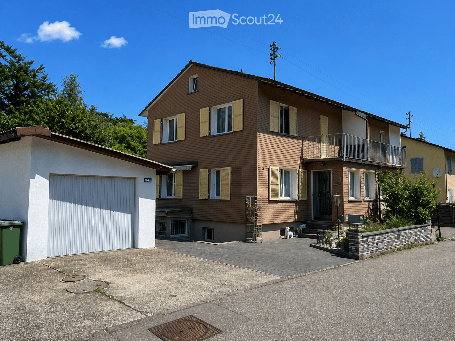 Two-story house with brown brick facade, white garage, and balcony on the upper floor. The house features multiple windows with shutters and a small garden area in front.