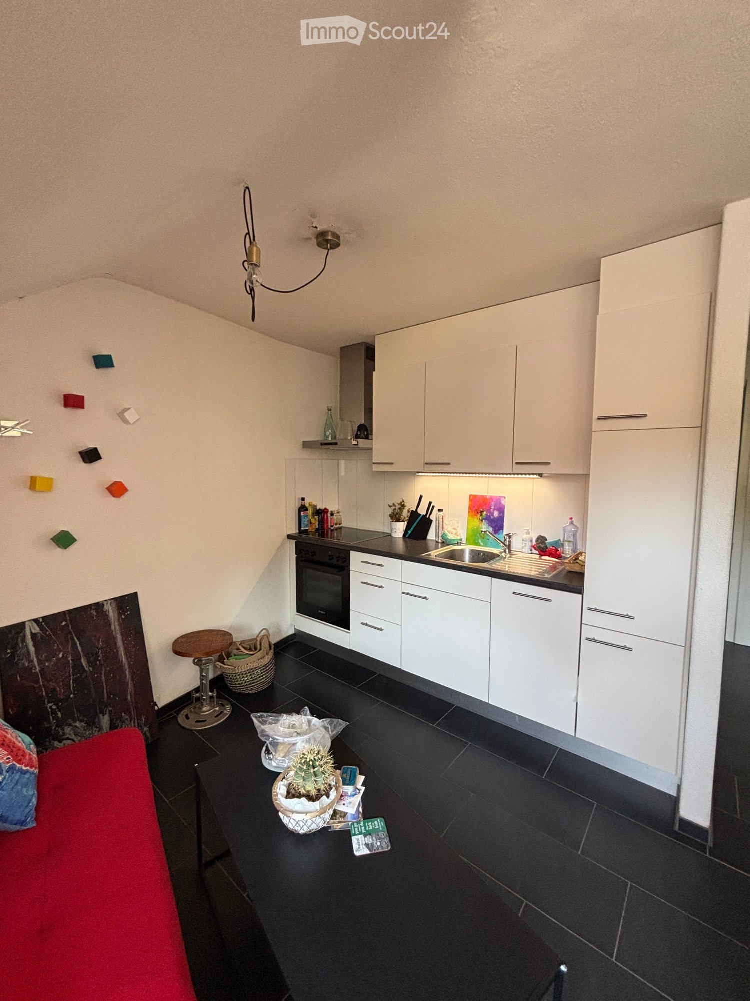 A modern kitchen with white cabinets, black countertops, and a small living area with a red couch and a small table. The kitchen features an oven, sink, and various small appliances. A cactus and a basket are placed on the floor near the living area.