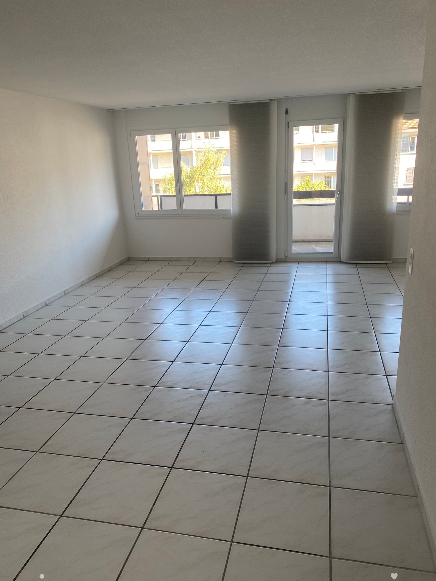 Empty room with tiled floor, white walls, and large windows with a sliding glass door leading to a balcony.