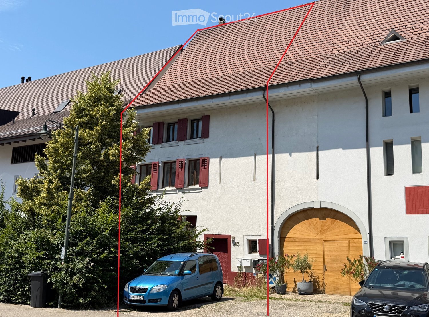 Two-story house with red-tiled roof, white walls, red shutters, and a wooden garage door. Two cars are parked in front.