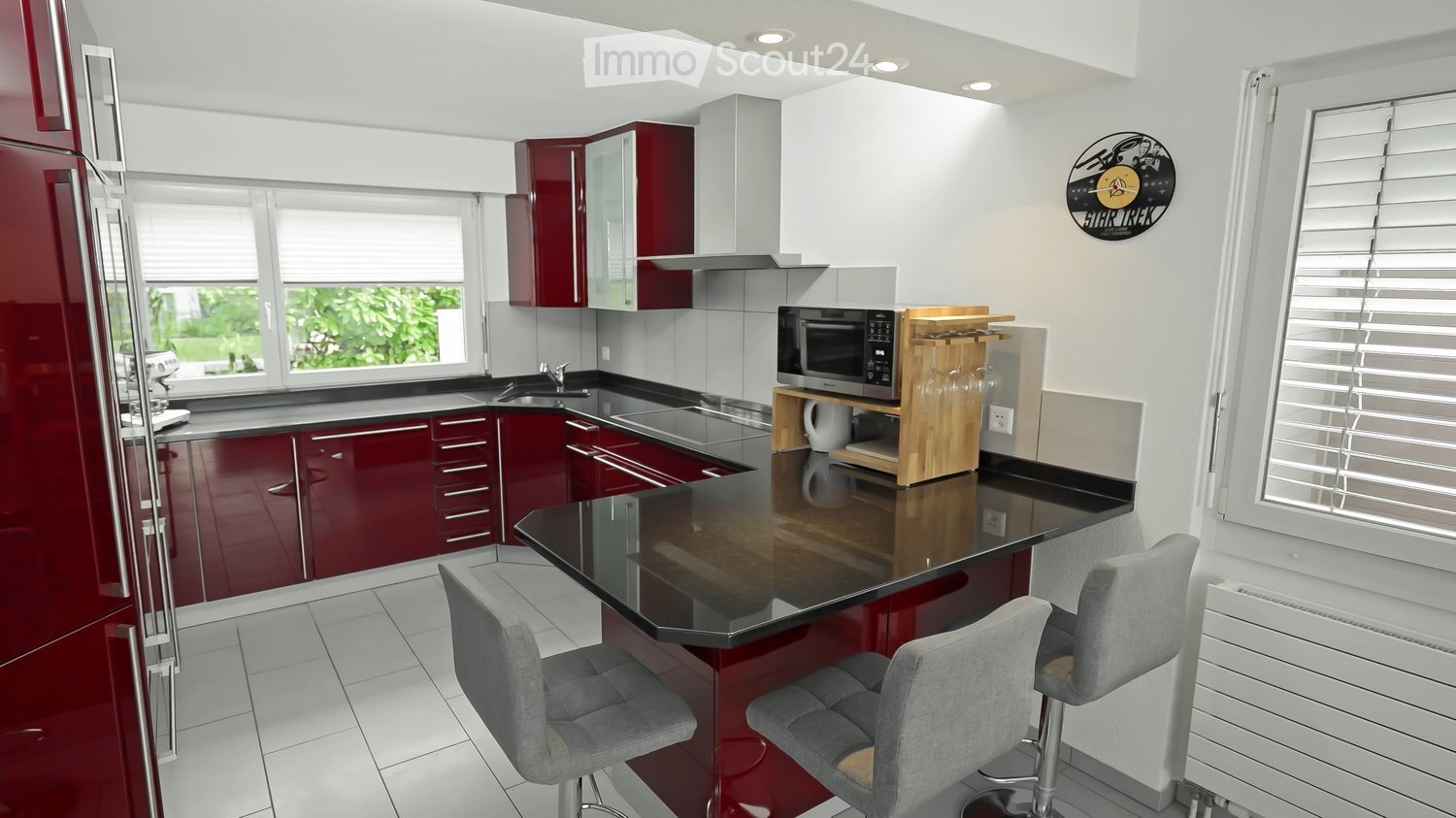 Modern kitchen with red cabinets, black countertops, stainless steel appliances, and a central island with bar stools.