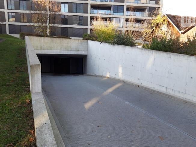 Modern multi-story building with a raised ground floor garage entrance, surrounded by greenery and a concrete pathway.