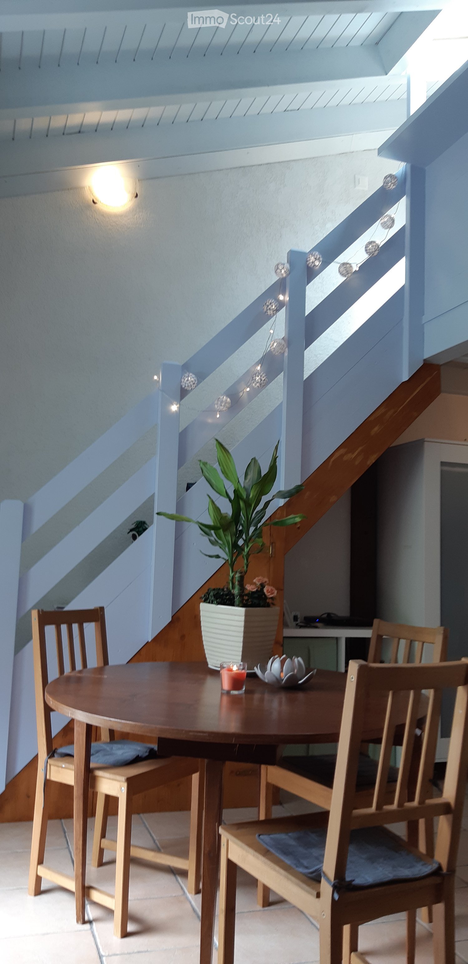 A small, well-lit dining area with a round wooden table, four wooden chairs, a potted plant, and a candle on the table. The area is adjacent to a staircase with a modern glass railing and string lights.