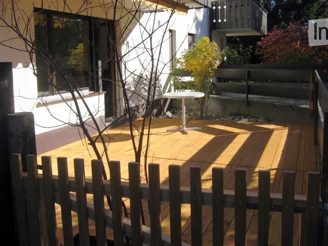 Wooden deck with a small round table, surrounded by a wooden fence, with a view of the house and garden.