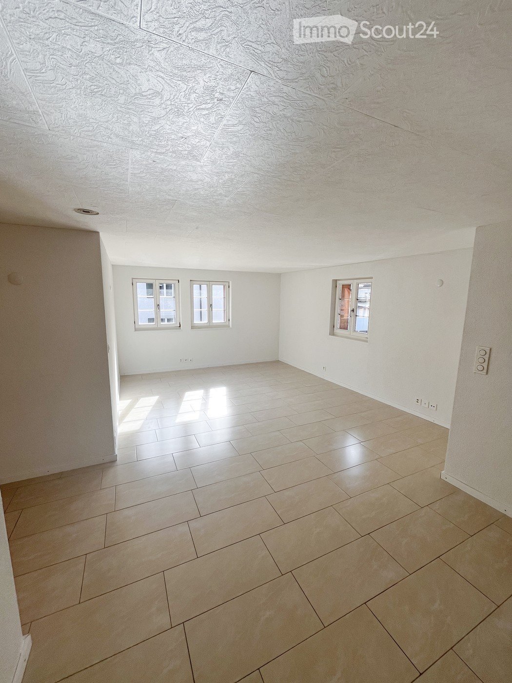 Empty room with light-colored tiled flooring and white walls, featuring three windows and a textured ceiling.