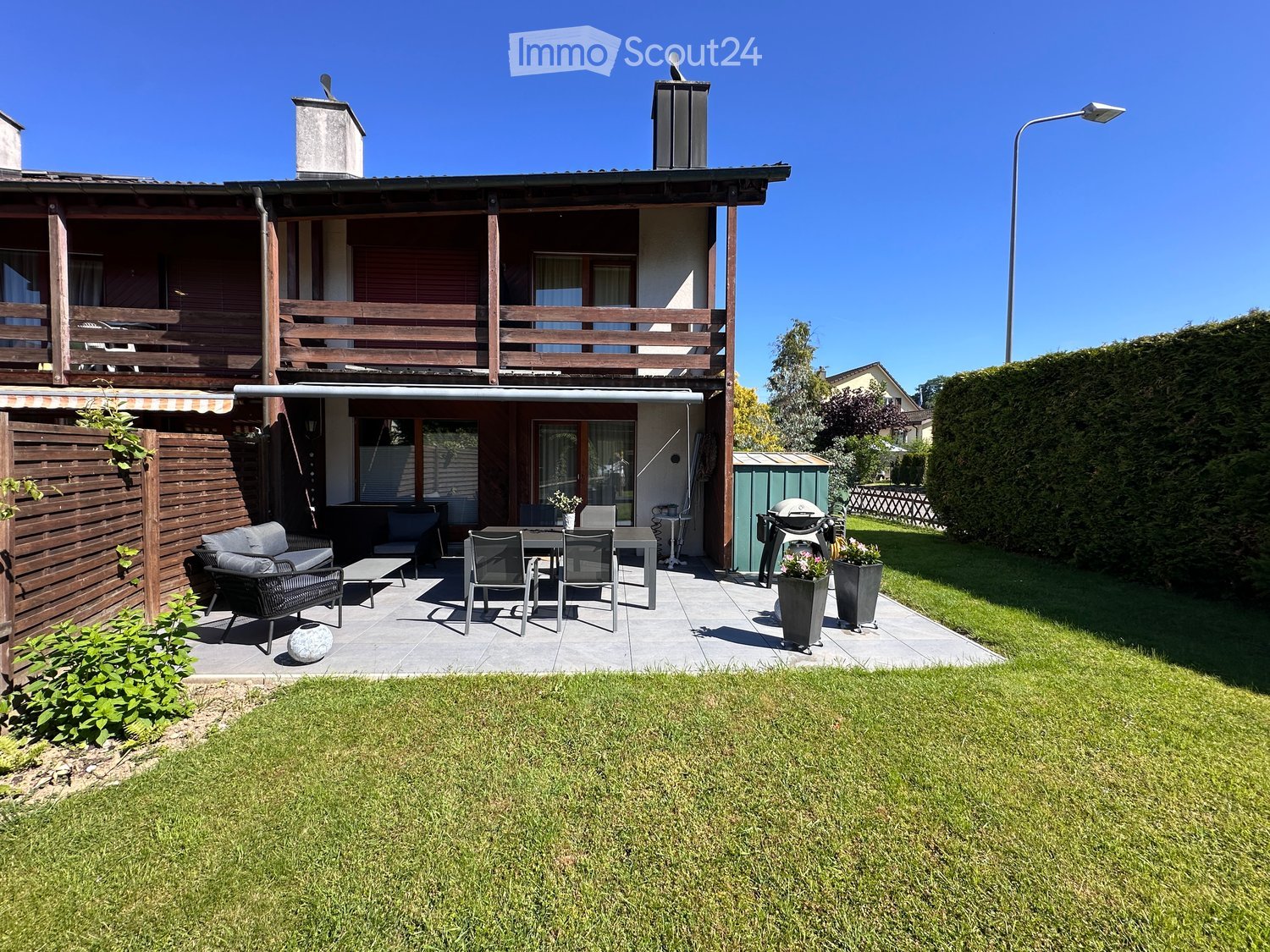 Well-maintained garden with a patio area featuring a sofa, table, chairs, and a grill. The house has a wooden balcony and a well-kept lawn.