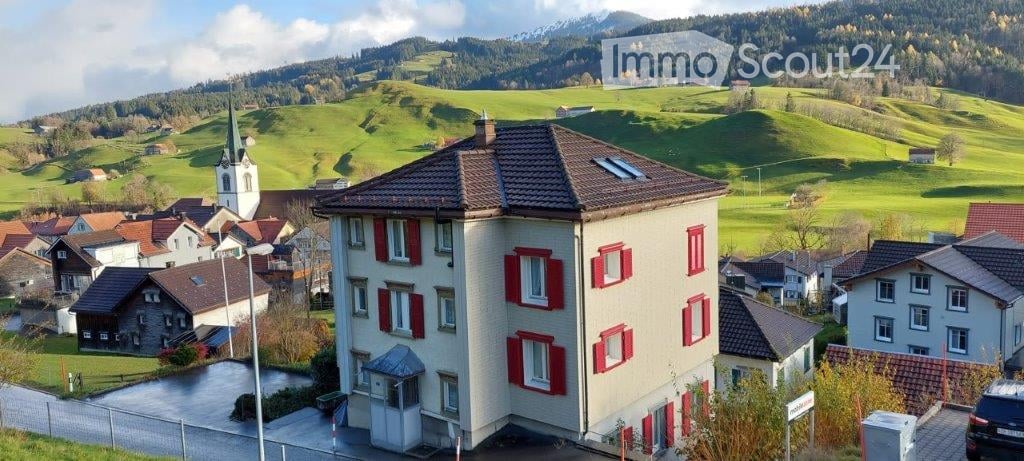 A multi-story residential building with red shutters and a tiled roof, situated in a picturesque village with green hills and a church in the background.