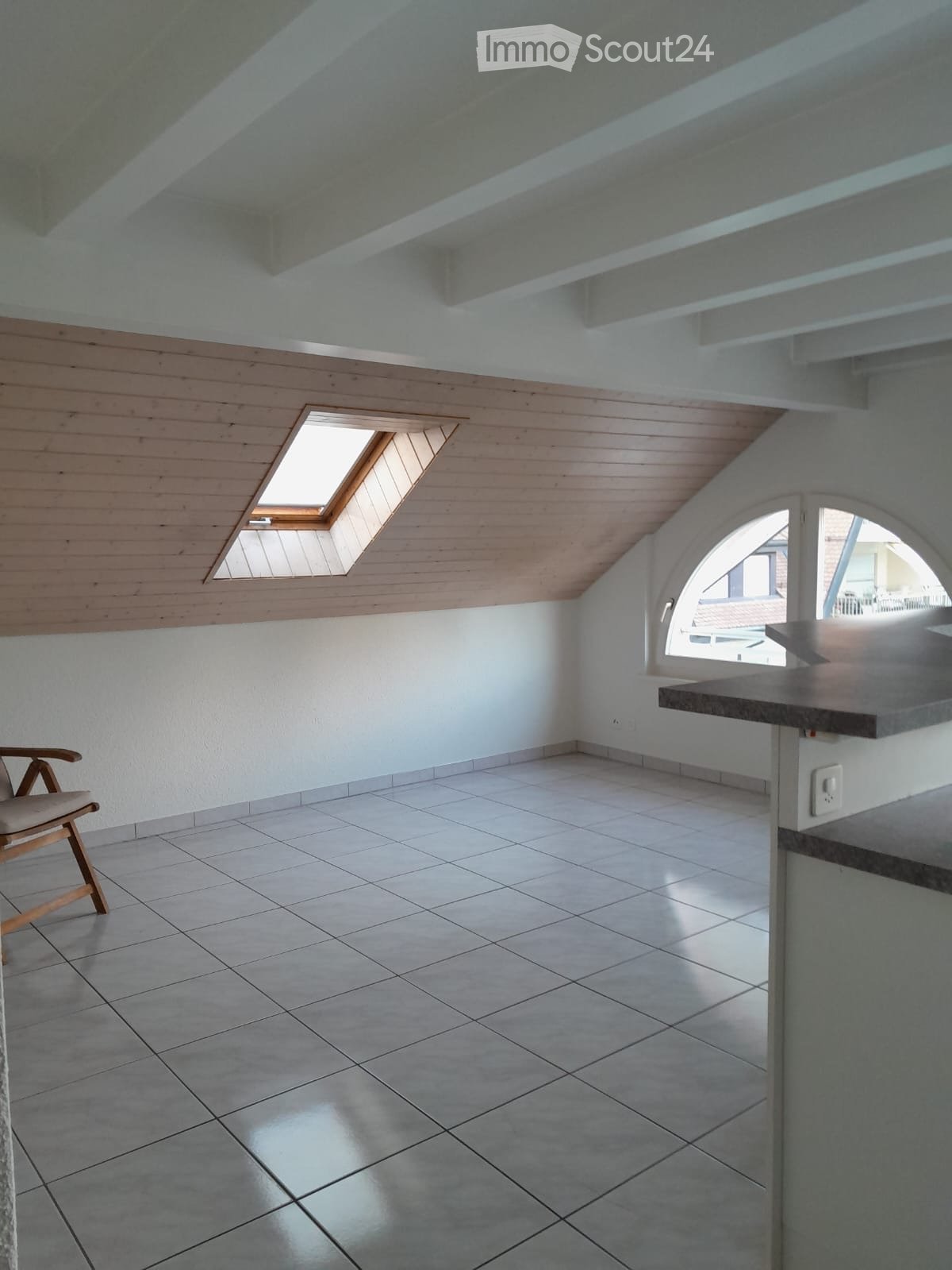 Bright, airy attic room with wooden slatted walls, skylight windows, tiled floor, and a single wooden chair near the wall.