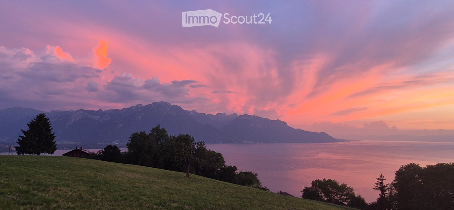 A scenic view of a lake and mountains at sunset, with a small house visible on the left side of the frame.