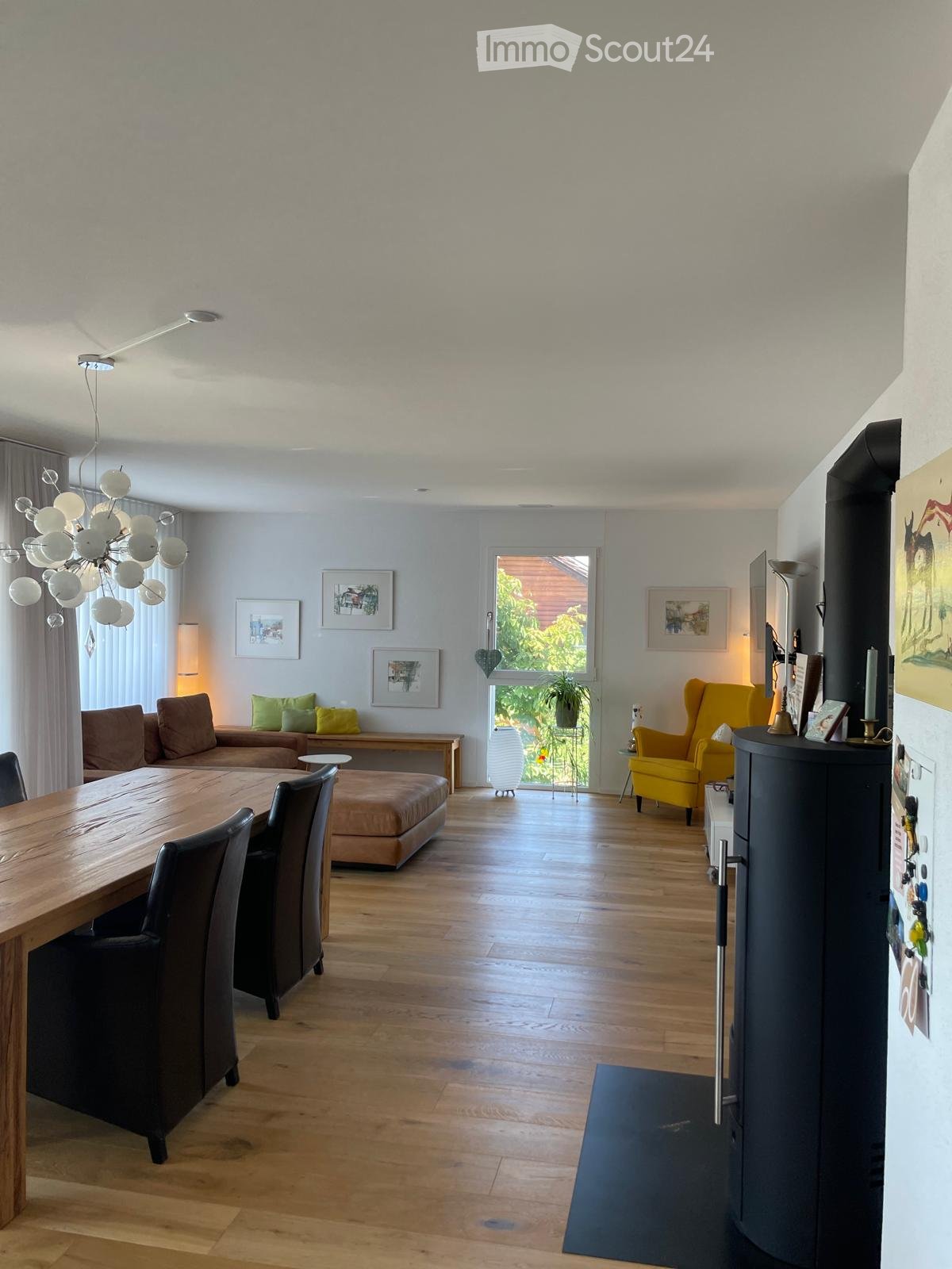 Open-plan living and dining area with wooden flooring, a large chandelier, a sofa, a dining table with chairs, and a yellow armchair.