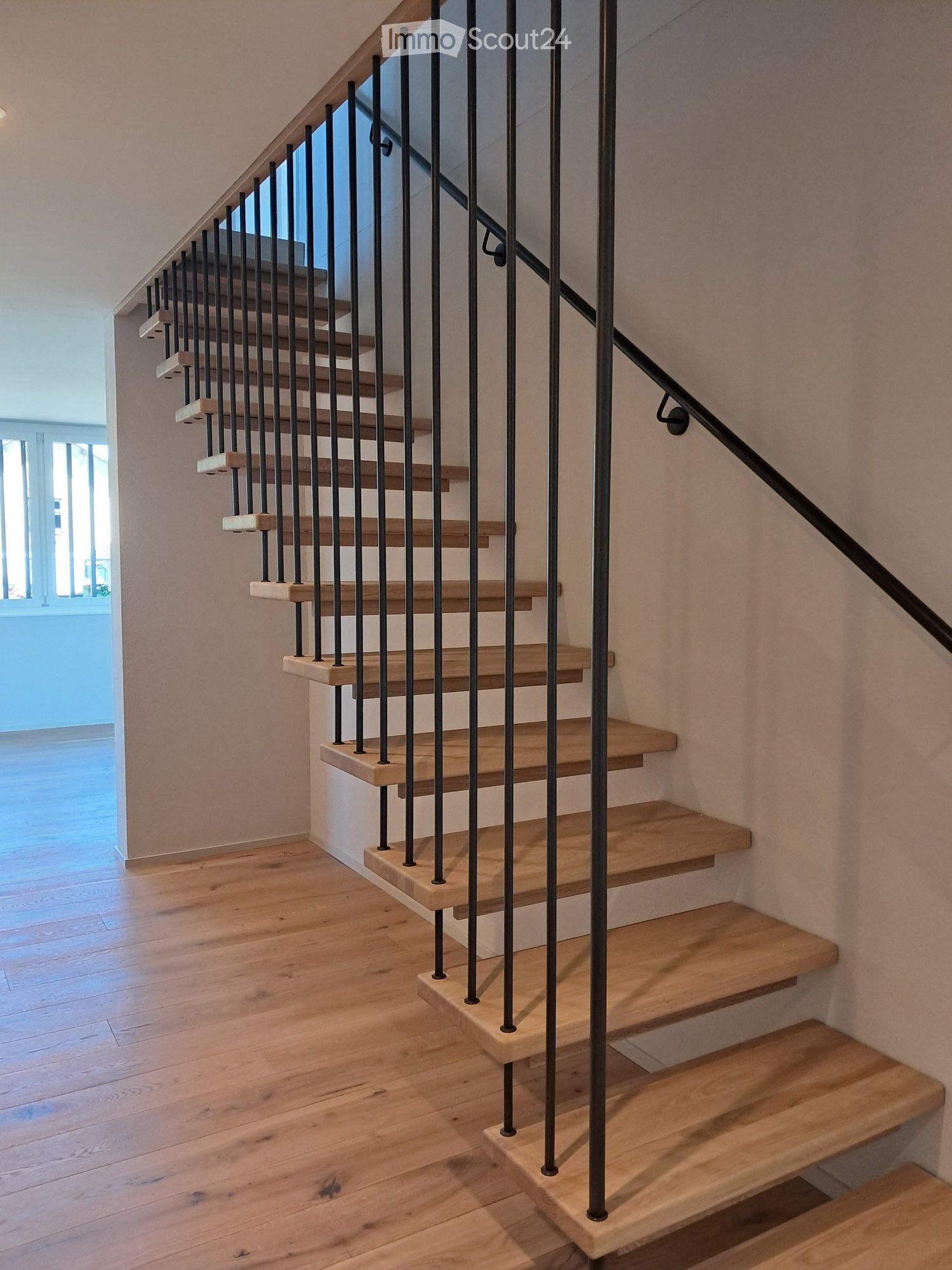 Wooden staircase with black metal balustrade, light wooden steps, and a handrail, set against light-colored walls and wooden flooring.