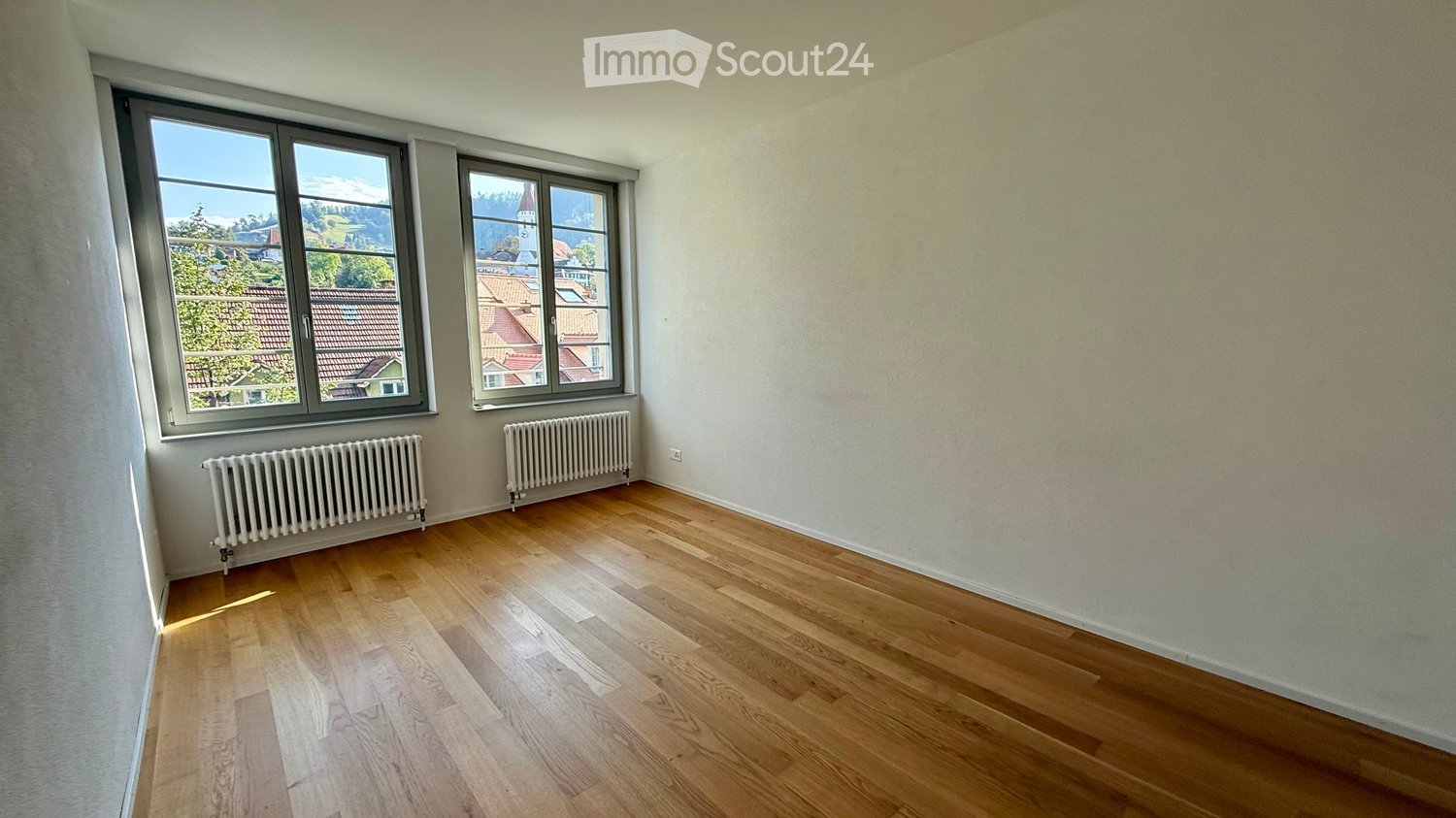 Empty room with wooden flooring, large windows offering a view of rooftops and trees, and radiators along the wall.