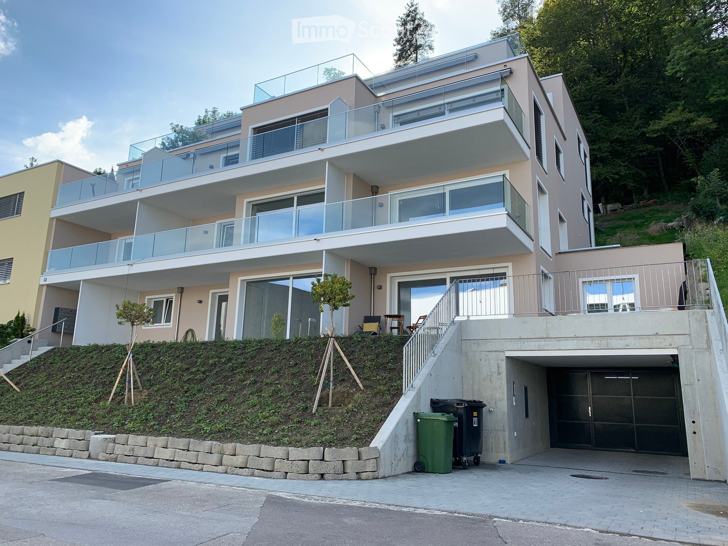 Modern two-story house with large glass balconies, raised ground floor, and garage. The house features a beige facade, multiple large windows, and a landscaped hillside entrance.