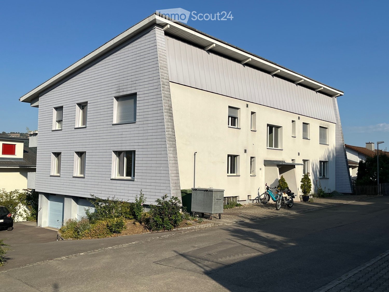 Modern two-story house with light grey siding, multiple windows, and a small garage. There are bicycles and a scooter parked near the entrance, along with some greenery and a trash bin.