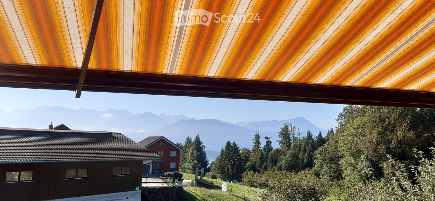 Balcony with a view of a house, trees, and mountains in the background.