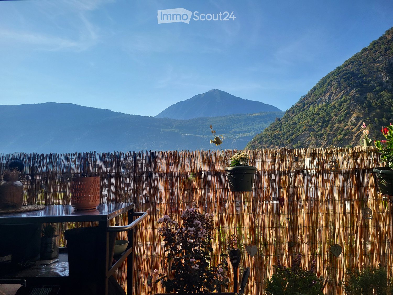 Balcony with a wooden privacy screen, overlooking a scenic mountain view. Features a small table with chairs, flower pots, and decorative vases.