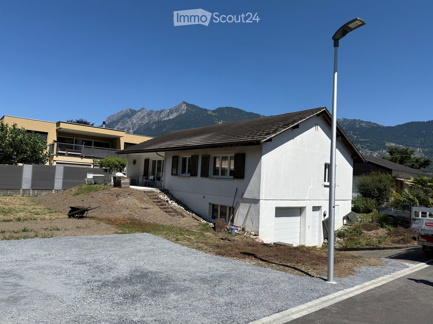 Single-story house with white exterior, multiple windows, garage, and raised ground floor. Located in a residential area with a view of mountains in the background.