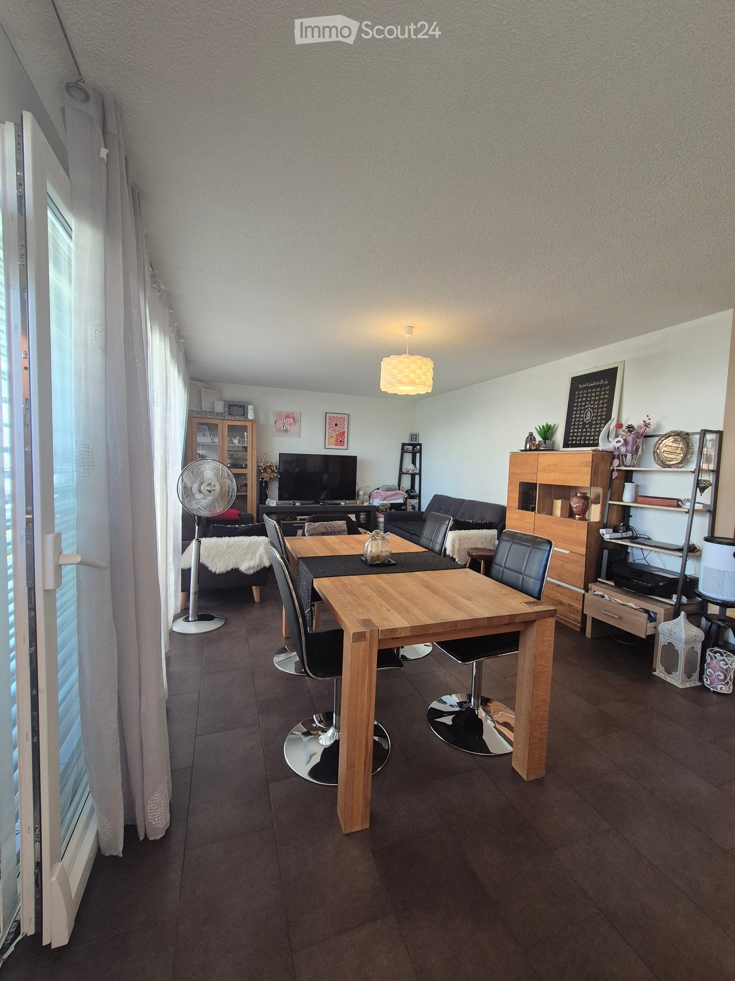 A modern living room with a wooden dining table, four chairs, a sofa, a fan, a television, and various decorative items on shelves and cabinets.