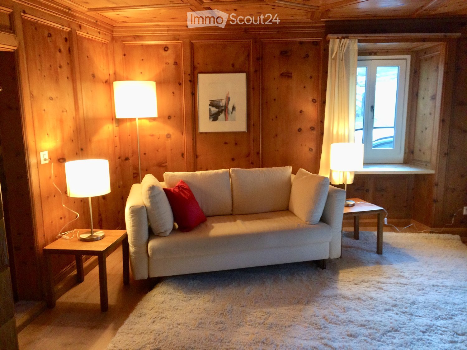 Cozy living room with wooden walls, a light-colored sofa with red throw pillows, two side tables with lamps, and a large picture frame on the wall.