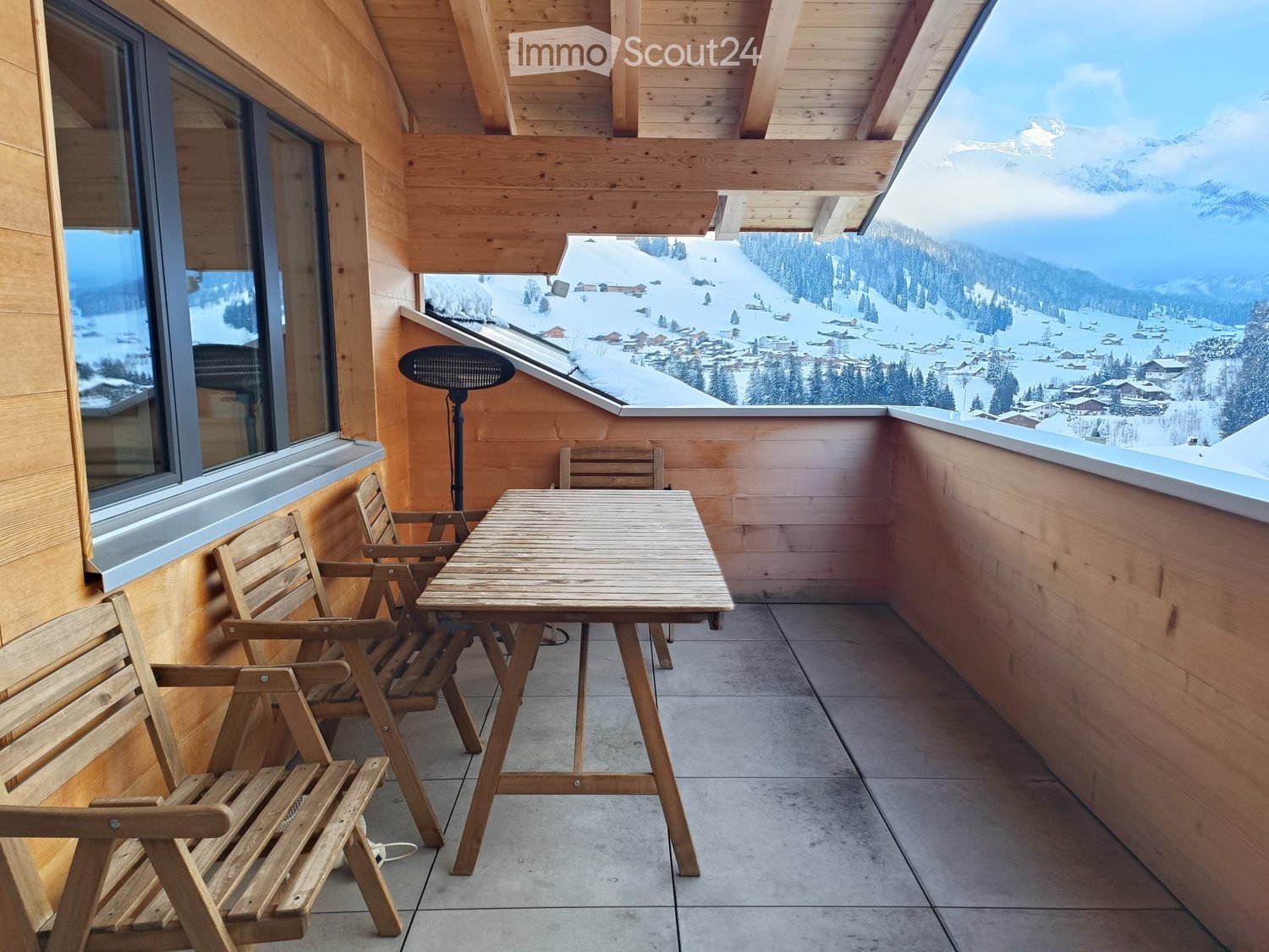 Wooden balcony with a table and chairs, overlooking a snowy mountain landscape.